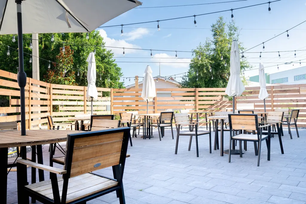 A sunny outdoor restaurant patio with wooden tables, white umbrellas, and string lights under a blue sky.