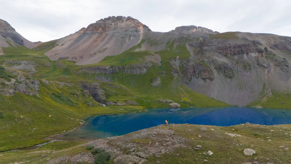 A vivid blue alpine lake beneath rugged Colorado peaks, with a lone hiker on the grassy shore.